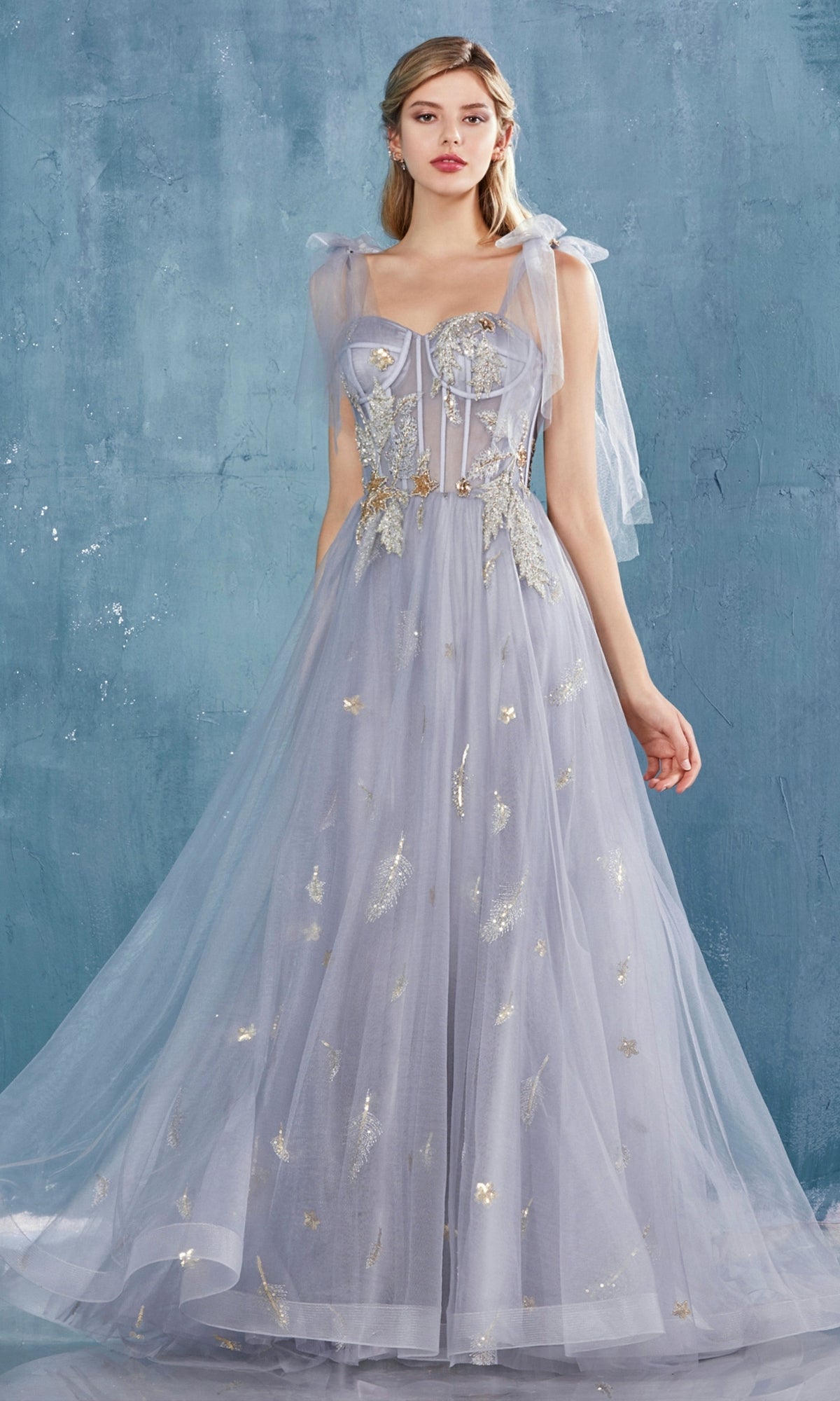 A woman poses against a blue textured wall wearing the Shoulder-Bow Long Corset Prom Ball Gown A0824, featuring lavender tulle, gold embellishments, a corset bodice, and sheer ribbon-tied shoulder straps.